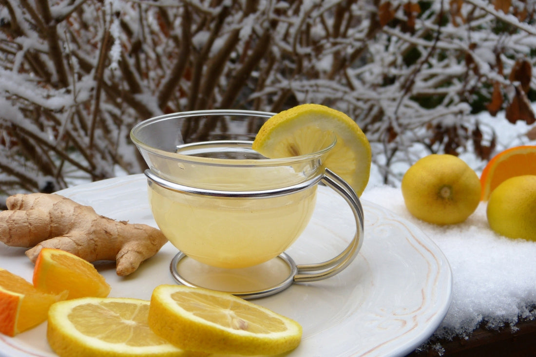 image of a glass cup containing herbal fruit tea standing on a white saucer or plate, garnish with a slice of lemon, and with sliced lemon and fresh ginger on the plate next to the cup. There is snow in the background.