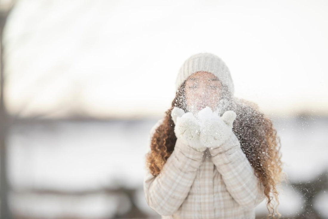 Image of a woman in winter wearing a while woolly hat scarf gloves and coat, outdoors in the snow, with long curly brown hair coming out under her hat. She is blowing snow from her hands towards the camera.