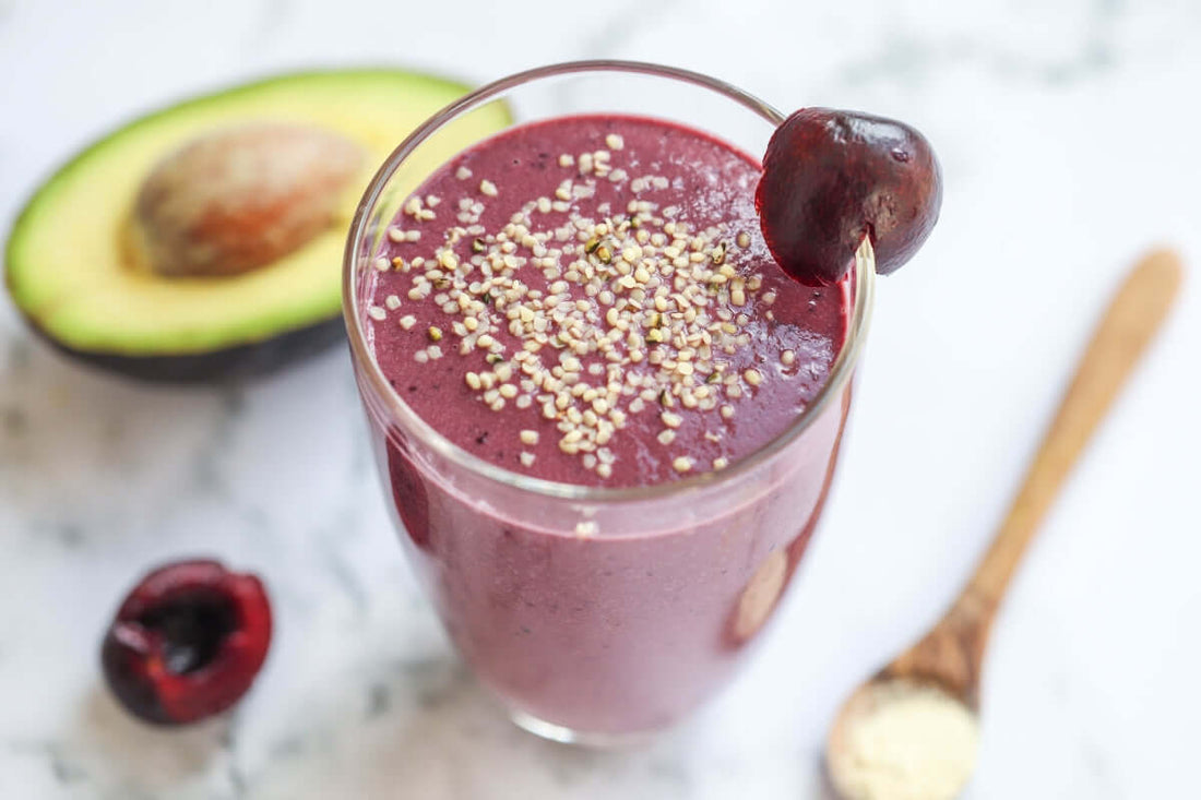 image of a purple black forest and avocado protein smoothie served in a clear glass with a cherry garnish and an avocado half to the side