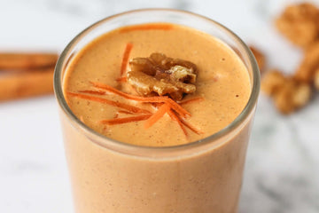image of an indulgent orange carrot cake smoothie served in a clear glass and topped with a walnut and grated carrot. There are cinnamon sticks and walnuts in the background