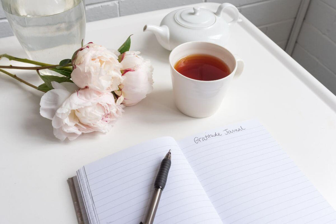 an open gratitude journal with white paper inside and a gold pen lying across it on a white desk with a cup of tea and pale pink roses and a glass of water