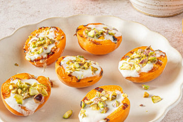 Grilled BBQ Apricot halves filled with yoghurt and topped with chopped pistachio nuts, on a white plate with scalloped edges on a white kitchen worktop