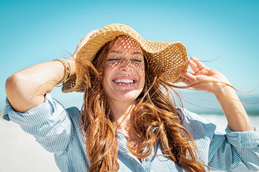 woman with long red hair and a wide brimmed summer hat laughing and holding her hat down with blue skies behind