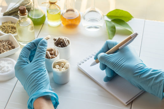 image of hands in blue latex gloves holding and inspecting several supplement capsules and liquids and tablets and making notes on a small notepad with a pen