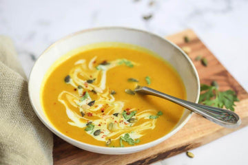 Curried Pumpkin & Butternut Squash Soup dressed with healthy seeds in a rustic white bowl with a spoon in it sitting on top of a board with herbs