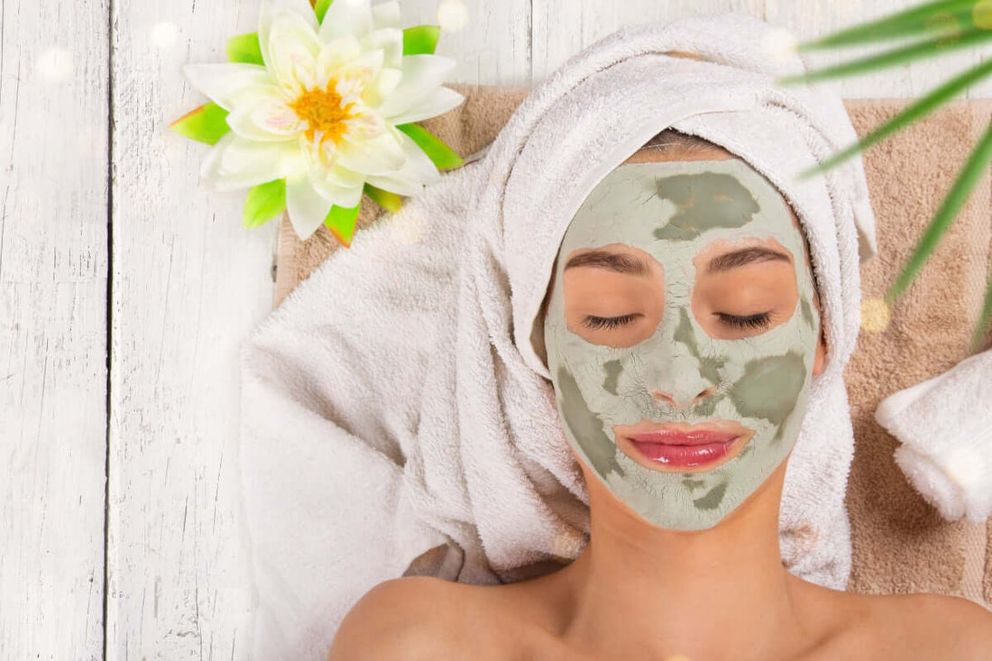 image taken from above of the head and shoulders of a woman lying down with a towel on her hair and a green clay face mask on. She is having beauty treatments in a spa setting and has a yellow flower to one side and a green leafy plant to the other side. 