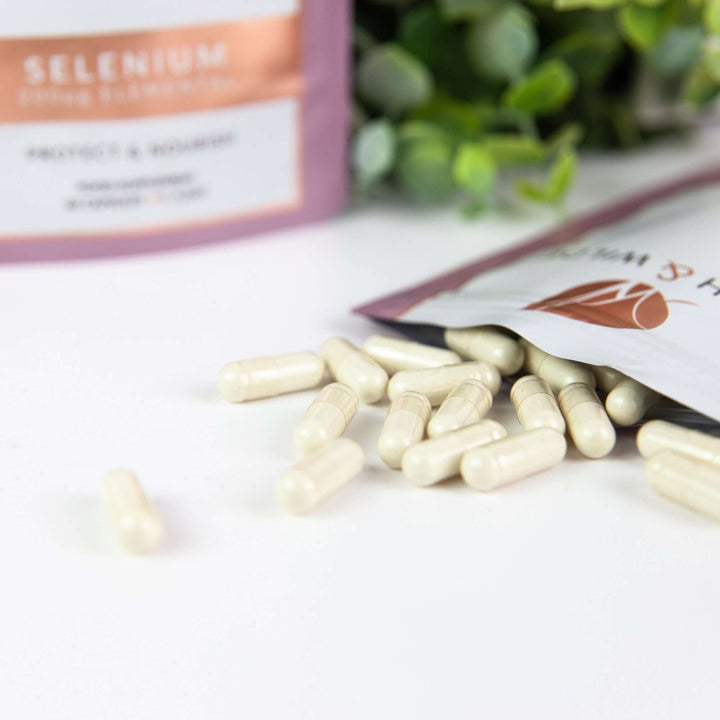 close up image of Birch & Wilde selenium capsule health supplement refill pouches. One pouch is standing up and the other is on its side with selenium capsules spilling out. There is a green plant in the background. 