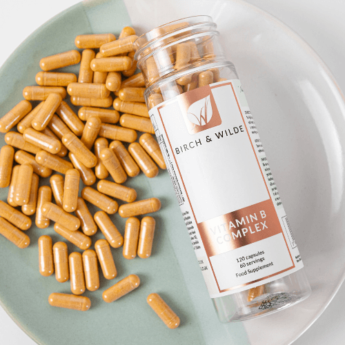 flat lay of of Birch & Wilde vitamin B complex bottle lying on a pale green and white plate with scattered yellow vitamin capsules filling the rest of the plate