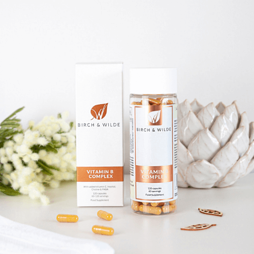  product lifestyle shot from front of Birch & Wilde Vitamin B complex capsules with scattered vitamin capsules and a white pot and green plant in the background