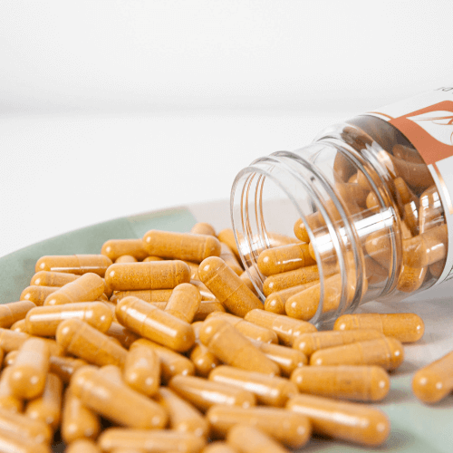 close up image of Birch & Wilde vitamin B complex capsules being poured from a bottle onto a pale green and white plate with scattered yellow vitamin capsules filling the plate