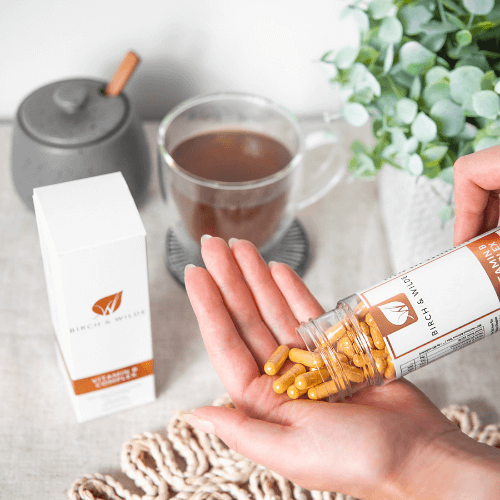 image of Birch & Wilde vitamin B complex capsules being poured from a bottle into a cupped hand below with a cup of tea and a green plant in a pot in the background