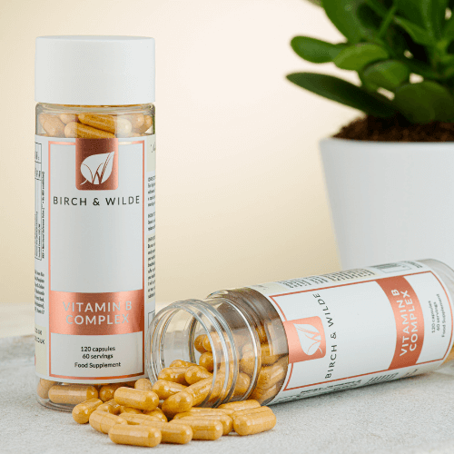 image taken from the front of two bottles of Birch & Wilde vitamin B complex capsules. one bottle is standing up and one bottle is lying on its side with yellow vitamin capsules spilling out. There is a green plant in a white pot in the background. 